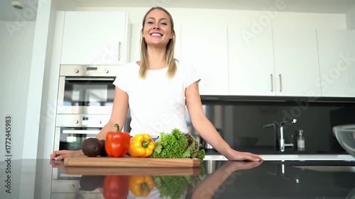 Smiling Young Woman In A White T Shirt Standing In A Modern Kitchen With Fresh Vegetables On A Countertop