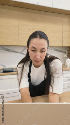 Young woman in black apron standing at kitchen counter and looking at laptop screen, concept of searching recipes and cooking ideas online at home