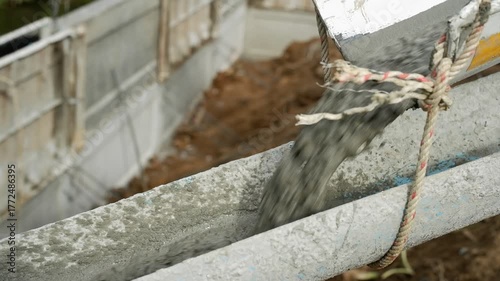 A dynamic, close-up shot captures the process of pouring fresh, wet concrete into a makeshift formwork channel on a construction site. The thick, viscous mixture is rapidly flowing, creating a motion 