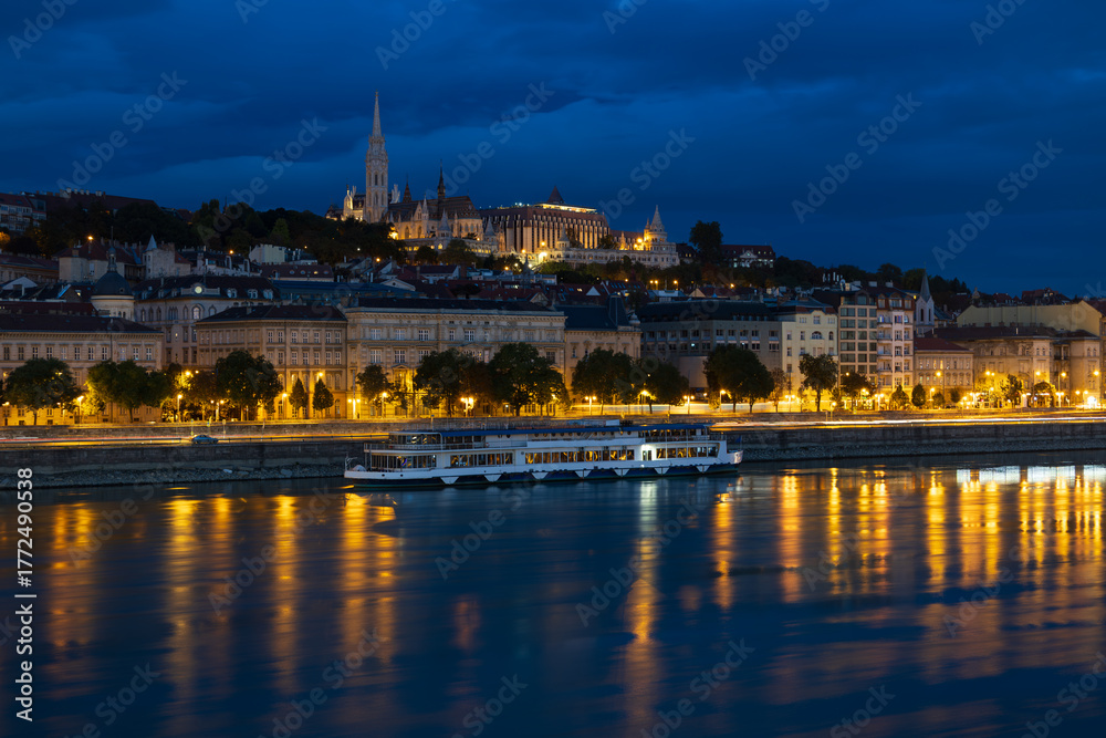 Fototapeta premium Blue hour view of boat on the Danube River, the coastline and the illuminated Buda Castle complex, Budapest, Hungary