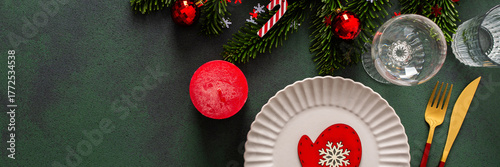A cozy Christmas table with an empty plate, cutlery and candle on a rich green background. Red Christmas tree decorations and spruse branches complete the festive look, banner