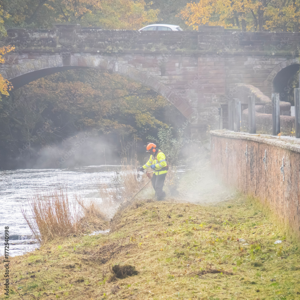Fototapeta premium Environmental agency worker cutting grass on a riverbank to protect from flooding.