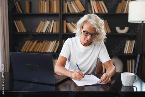 Middle aged senior woman using laptop computer writing notes at workplace. Focused mature old businesswoman work at office making written records doing online work. Mature boss professional worker