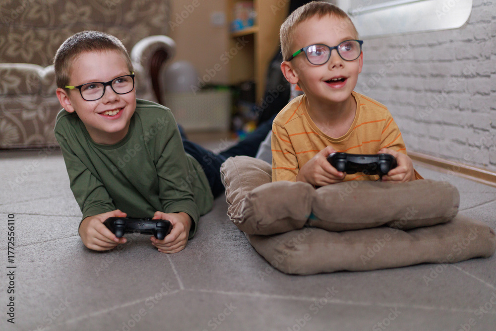 Fototapeta premium Happy children lying on cushions and playing video game together at home. Concept of friendship, joy, and teamwork in childhood with copy space.