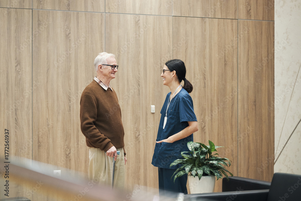 Fototapeta premium Senior Caucasian man standing and talking with young adult Caucasian female nurse in medical uniform in modern healthcare facility, both smiling and making eye contact
