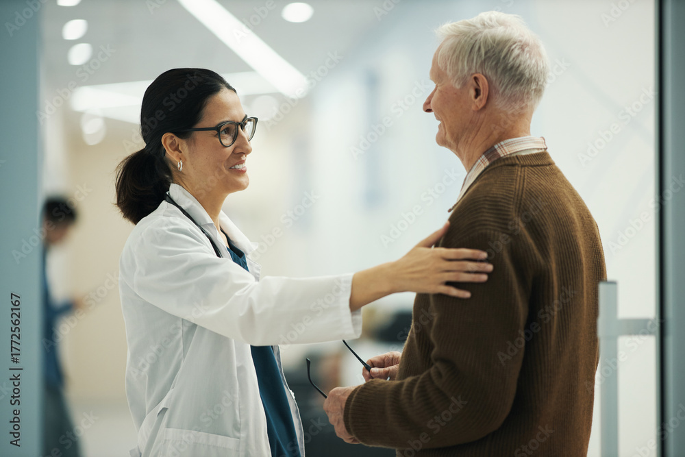 Obraz premium Caucasian female doctor greeting senior Caucasian male patient in medical facility, doctor smiling and placing hand on patients shoulder, while holding eyeglasses in other hand