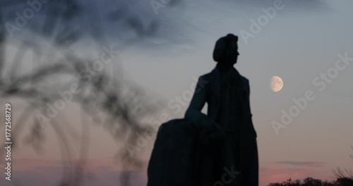 Silhouetted statue against a pastel dusk sky as a waxing gibbous moon rises. Out-of-focus branches frame the scene, evoking solitude, memory, and quiet contemplation.