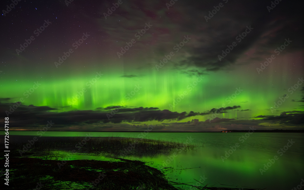 Obraz premium Vivid northern lights glowing green and purple over a calm lake with reflections and scattered clouds under a starry night sky.