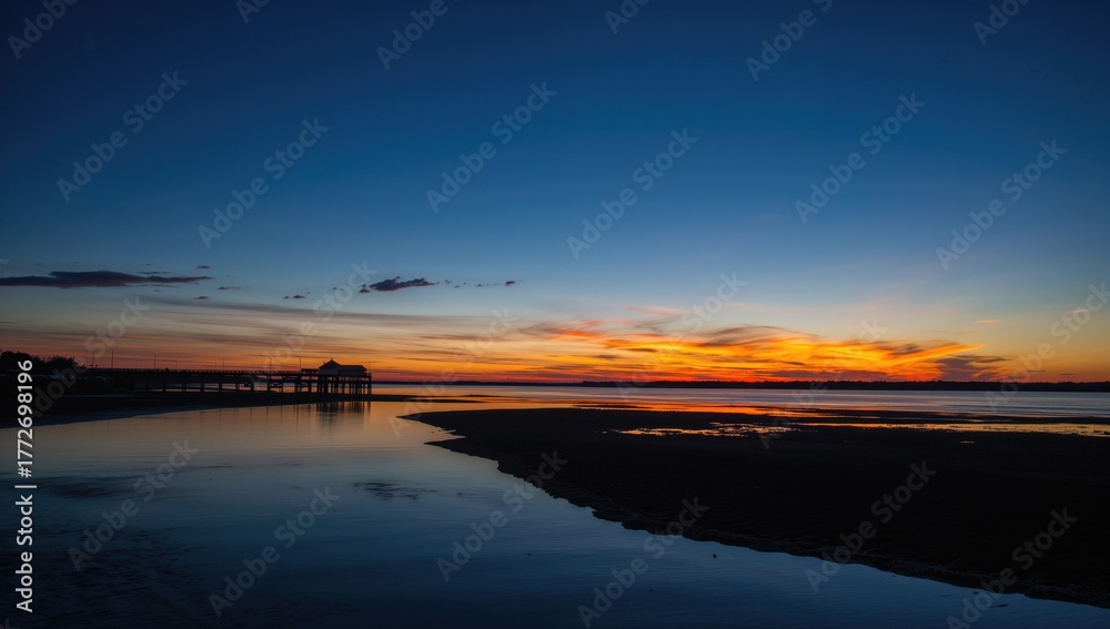 Obraz premium Tranquil river shore at dusk featuring a darkened pier, still water mirroring the warm colors of the evening sky, and a serene sandy area.