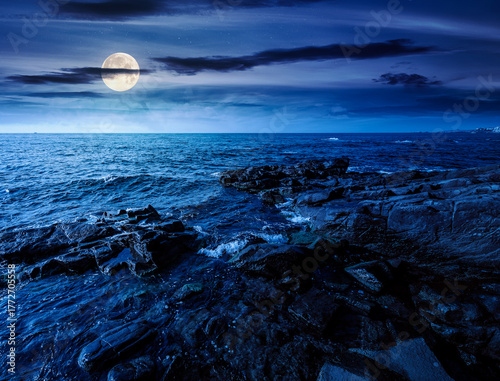 night landscape of rocky sea coast. beautiful full moon over horizon. summer coastline scenic view. composite scene during midnight. magical seascape vacations. waves and boulders in the dark