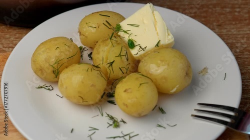 Close-up: on a white plate lies hot boiled potato sprinkled with fresh dill. A piece of butter slowly melts on top, dripping down the potato. A fork rests nearby video is out of focus