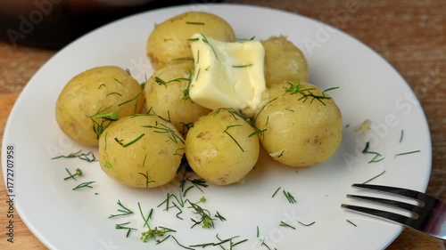Close-up: on a white plate lies hot boiled potato sprinkled with fresh dill. A piece of butter slowly melts on top, dripping down the potato. A fork rests nearby. Homemade food, warmth, and comfort.