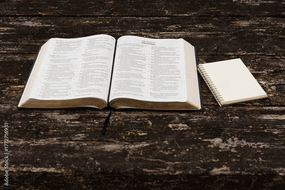 Obraz premium Flat lay of open Holy Bible (Psalms 149-150, Proverbs 1) and notepad on rustic wood table. Top view in natural daylight. Symbolizes spiritual study, wisdom, reflection, and devotion.