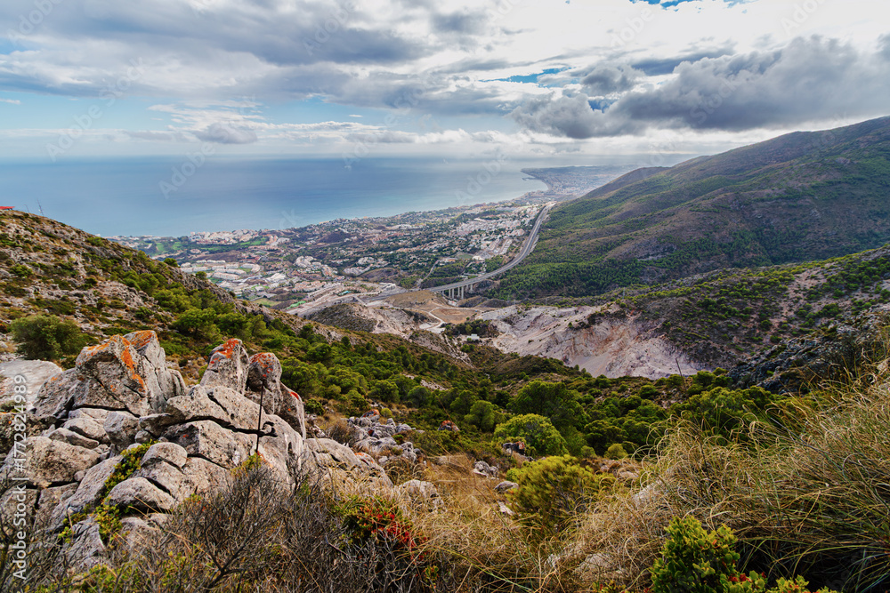 Fototapeta premium Spain - From a rocky mountain vantage point, a coastal city and highway stretch along the ocean under a dramatic cloudy sky.