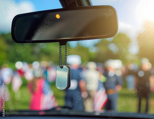 Car rearview mirror with military dog tag hanging, memorial event in background.