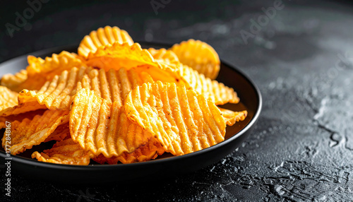 Plate of golden ridged potato chips on textured black surface. Tasty snack. Fast food.
