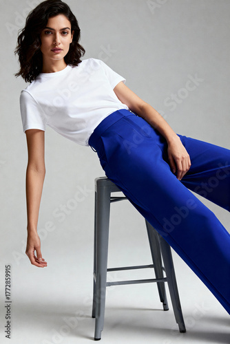 A stylish female model with dark wavy hair poses on a stool in a basic white t-shirt and vibrant blue wide-leg trousers against a gray studio background.