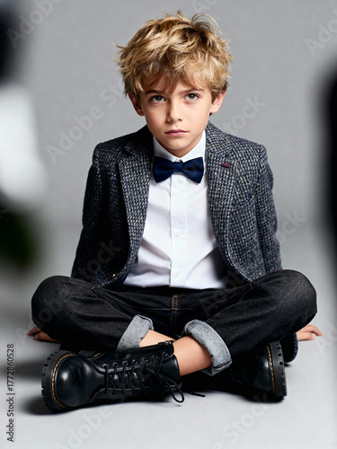 A stylish young boy with blond hair, wearing a tweed blazer, bow tie, and boots, sits cross-legged against a simple gray studio background.