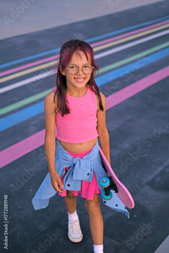 A beautiful girl in stylish pink clothes holds a skateboard in her hands. A child on the playground of a skate park