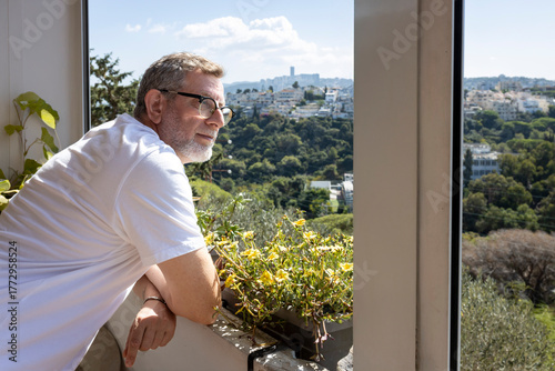 Fotografie A man in a white t-shirt and glasses stands on a balcony, looking out over a hillside covered in lush greenery and a distant cityscape under a bright, sunny sky