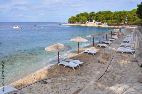 View of golden sand meeting turquoise water under a clear blue sky, framed by a rustic lone thatched parasols in a row. Pebbles beach. Tropic place in Croatia shoreline. Kvarner bay on Adriatic Sea