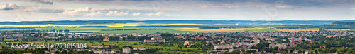 A high-angle panoramic view of the town of Zolochiv in Lviv Oblast, Ukraine. The cityscape, featuring a railway and red-roofed houses, is surrounded by green hills and agricultural fields.