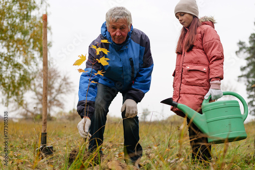 An adult and a child work together to plant a small tree in a grassy field as they prepare for a colder season with falling leaves