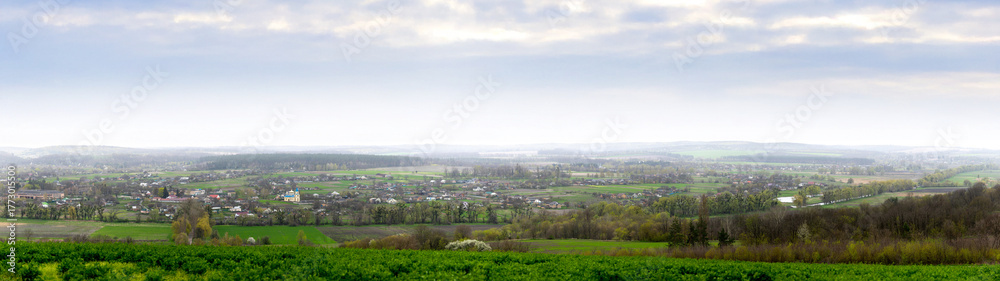 Obraz premium A panoramic view of Mliyiv village, Cherkasy region, Ukraine, in the Vilshanka River valley, during spring late April. The photo shows houses and buildings surrounded by green fields and rolling hills