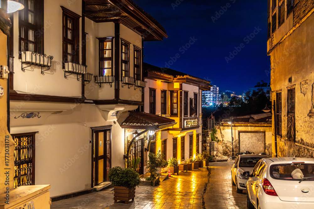 Fototapeta premium A narrow street in Kaleici old town illuminated by evening lights, featuring traditional Ottoman houses with cumba balconies, flower pots, and reflections on wet cobblestones. Antalya, Turkey.