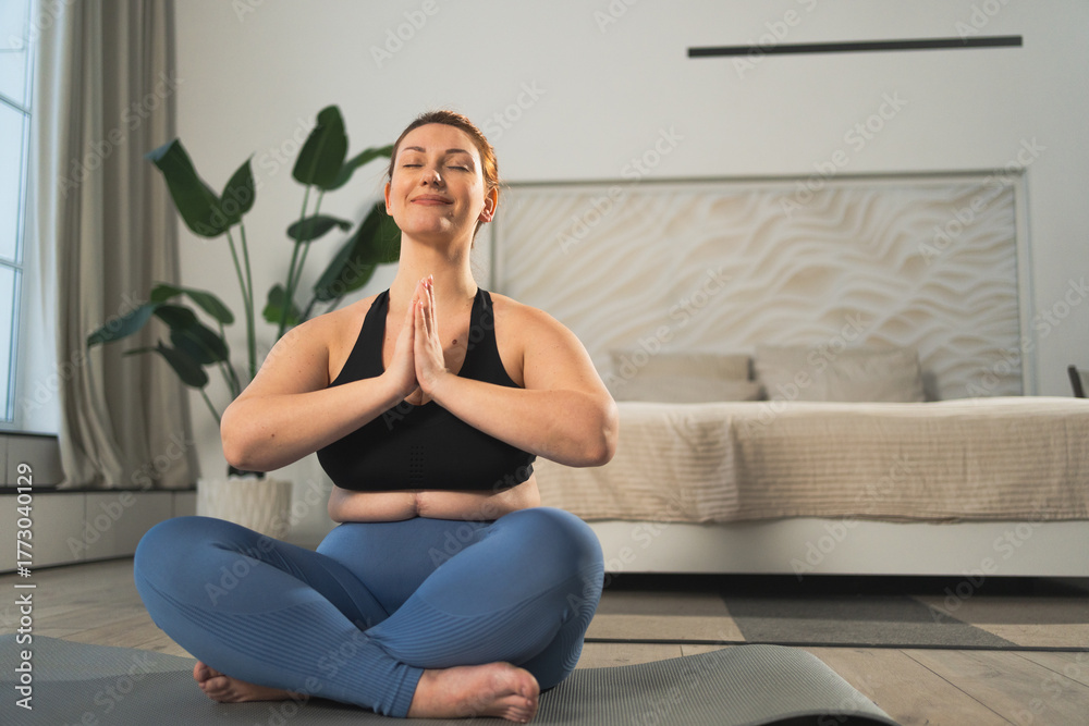 Obraz premium Yoga mindfulness meditation. Woman practicing yoga in living room at home. Plus size female sitting in lotus pose meditating smiling relaxing indoor. Girl doing breathing practice. Yoga at home
