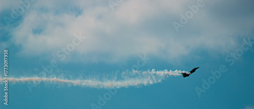 A fighter plane is flying in the cloudy sky with a trail of smoke that curls behind it. The wide perspective impressively reflects the elegance of flight and the vastness of the sky.