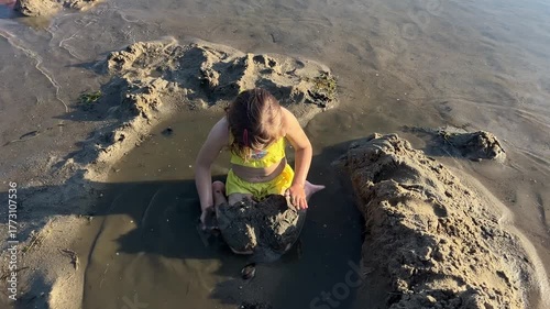 A girl in a yellow two-piece swimsuit plays in the sand on the river.