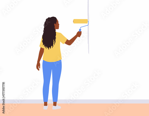 A woman with long dark hair paints a white wall with a yellow paint roller, viewed from her back.