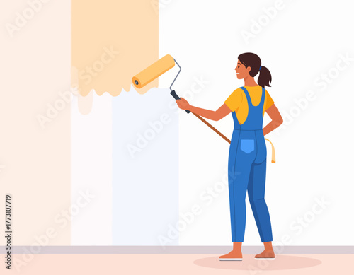 Woman in blue overalls painting a wall with a roller, applying yellow paint to the upper section, viewed from the side.