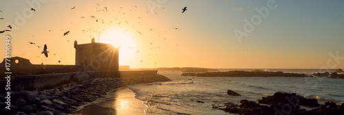 The sun sets behind a historic building in Essaouira, Morocco, casting a warm glow on the beach. Birds soar above the gentle waves, creating a serene coastal scene