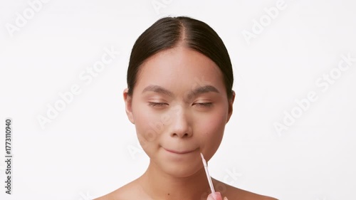 A beautiful young woman is pampering herself with skin care, applying lip gloss with focus and elegance, set against a clean background that enhances her serenity.