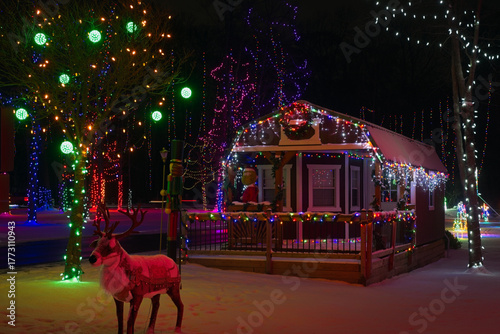 A reindeer bathed in red light stands before a small city park cottage as part of an extravagant Christmas lighting display.