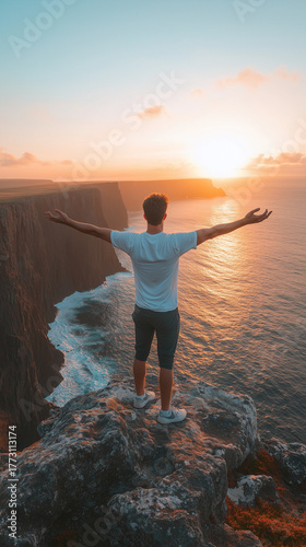 A person with outstretched arms stands on a high cliff overlooking the ocean, enjoying the magnificent sunset view.