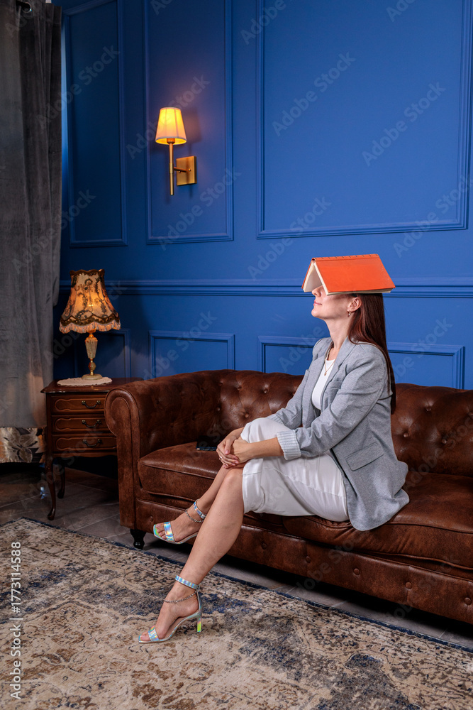 Fototapeta premium Woman covering her face with a book while resting on a luxurious leather sofa, depicting study break or mental exhaustion
