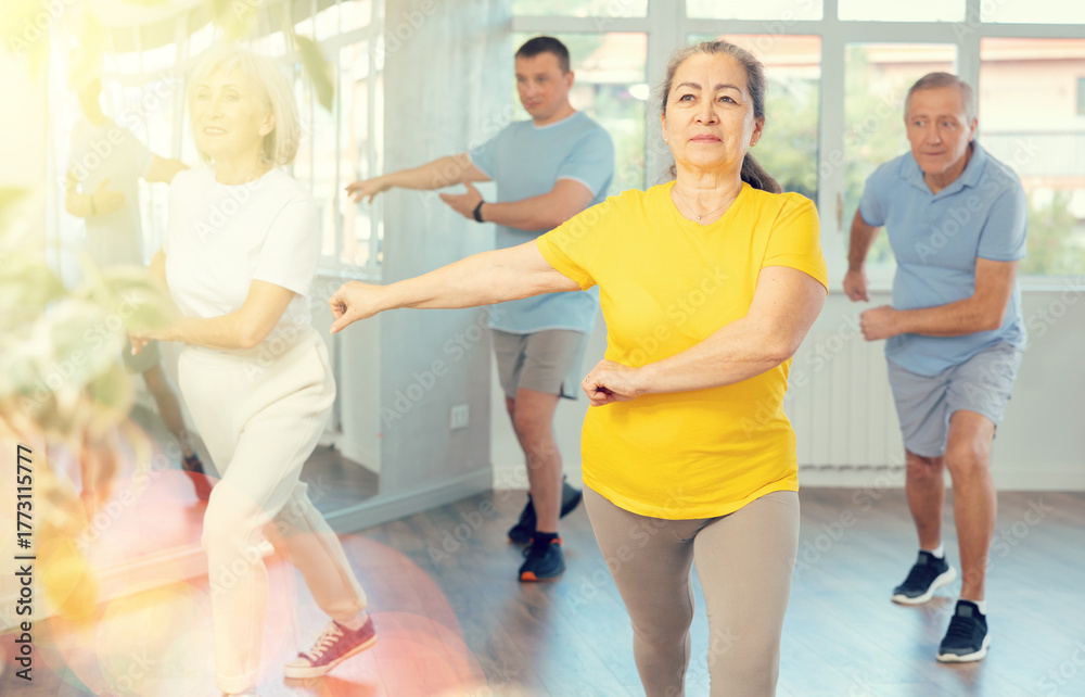 Obraz premium Positive elderly woman practicing Tai Chi with group of aged people, promoting health and wellness in bright training room