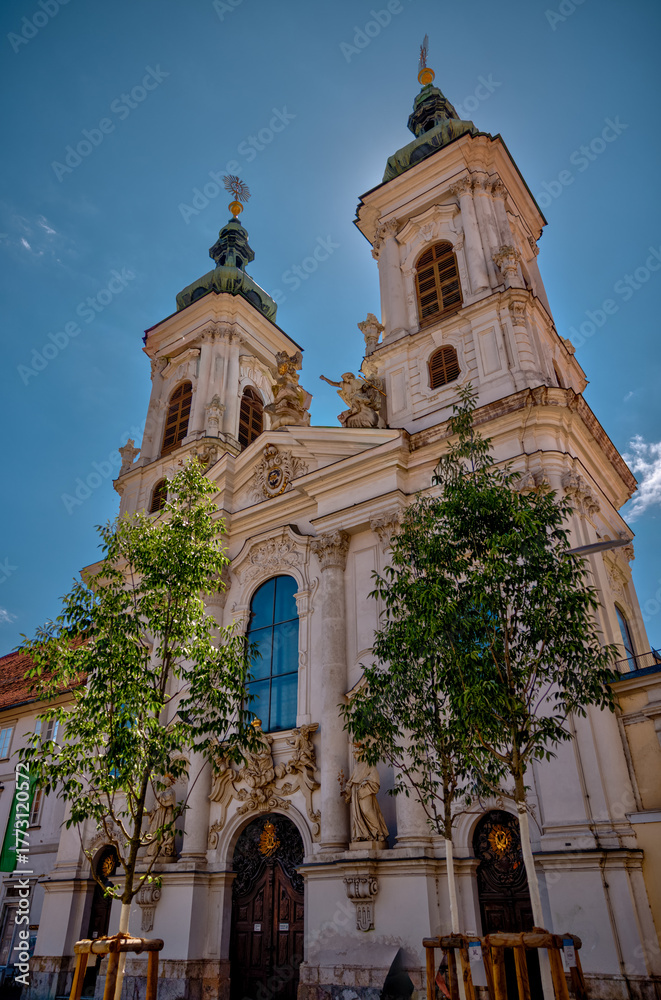 Fototapeta premium Graz, Austria – July 22, 2025: Exterior view of the Mariahilferkirche, a prominent Baroque-style Catholic church in Graz featuring twin towers and ornate statues on the façade. 