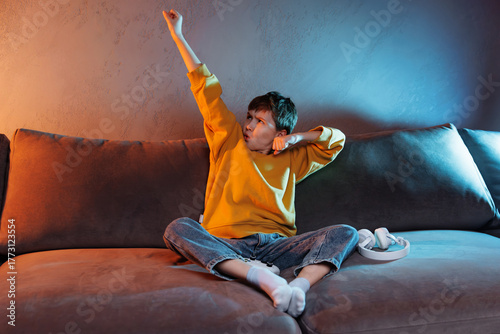 Young child in cozy yellow sweater sits on a sofa, striking a heroic pose with a determined expression, showcasing playful imagination and creativity in a vibrant living room setting