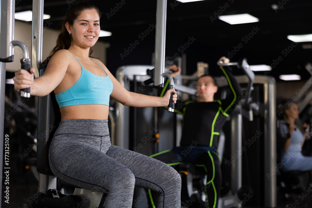 Fototapeta premium Concentrated young girl leading healthy active lifestyle doing strength training in gym, performing butterfly exercise for chest muscles in pec deck machine ..