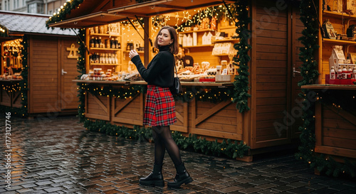 Stylish young woman in a plaid skirt visiting a festive outdoor Christmas market at night.