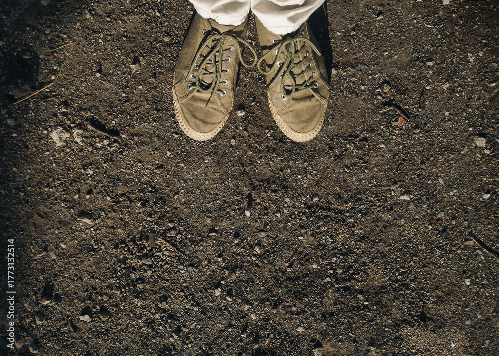 Obraz premium Khaki-colored moccasins with light soles on a man's feet in white pants against a gray dirt road with small stones. Concept of walking and relaxing in the fresh air.