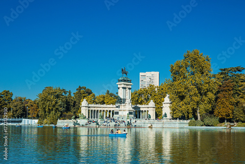 A calm lake reflects the grand monument at El Retiro Park in Madrid. People relax on steps and row small boats under a clear blue sky, surrounded by trees in warm seasonal colors.
