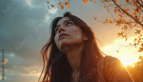 Young lady looks up to heaven at golden hour. Woman with long hair enjoys sunlight near tree with yellow leaves. Brunette dreams looks hopefully to sky at sunset time outdoors.