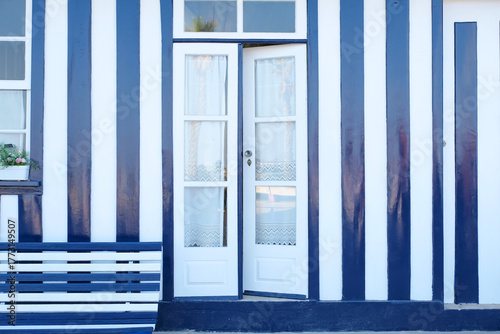 a blue and white striped house facade with a wooden bench and a open door, decorated with flower pots, Costa Nova, Aveiro, Portugal