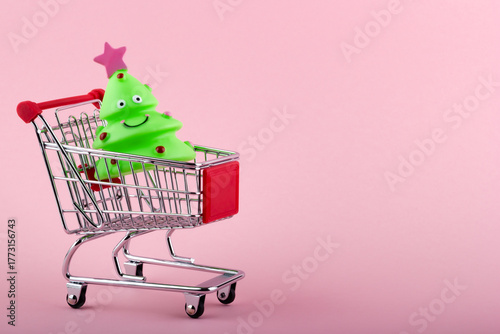 Grocery cart with small green toy Christmas tree on pink background. New Year shopping concept.