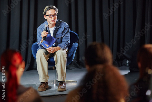 Valokuva Young adult man sitting on stage holding microphone and notebook, speaking to au